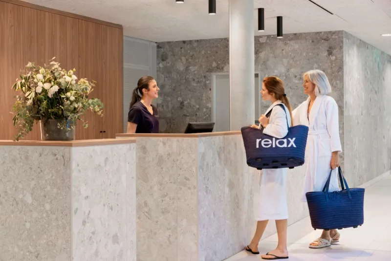 Eine Mutter und ihre Tochter in einer Hotellobby, in weißen Bademänteln und mit  Relax Taschen, lächeln eine freundlich aussehende Rezeptionistin an.